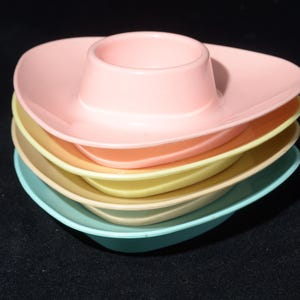 May include: A stack of pastel-coloured egg cups. The egg cups are in shades of pink, orange, yellow, and blue. Each cup has a circular centre for the egg and a wide, oval-shaped rim. The egg cups are stacked on top of each other.