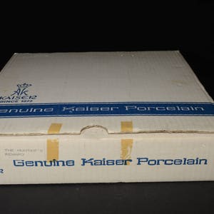 May include: A white cardboard box with blue and gold accents. The box has the text "Genuine Kaiser Porcelain" printed on it. The box is for a set of porcelain dishes.
