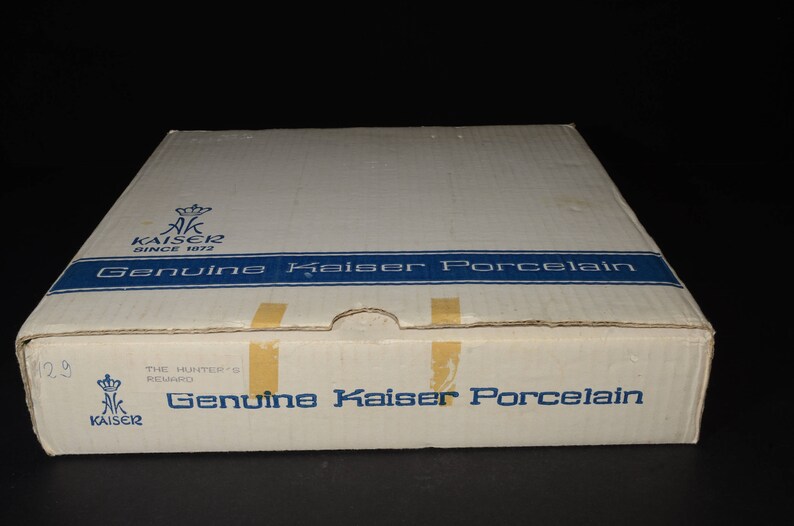 May include: A white cardboard box with blue and gold accents. The box has the text "Genuine Kaiser Porcelain" printed on it. The box is for a set of porcelain dishes.