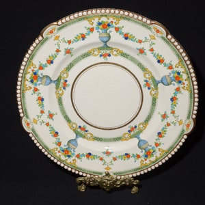 Pode incluir: Um prato de jantar de cerâmica branca com uma borda dourada e um design floral em tons de azul, verde, laranja e amarelo. O prato tem uma borda de contas e está em um suporte de metal dourado.