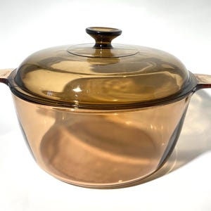 May include: A round, amber-coloured glass cooking pot with a matching transparent amber glass lid. The pot features two integrated side handles and a central knob on the lid. The glass is translucent, allowing light to pass through the vessel.