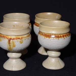 May include: Set of four ceramic goblets with a white base and brown and yellow glaze. The goblets have a textured surface and a rounded bowl.
