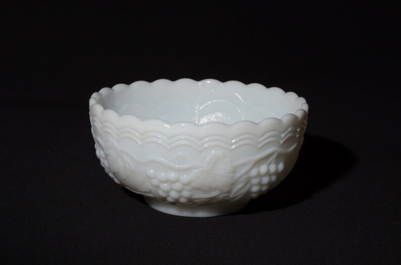 K&ouml;nnte beinhalten: Eine wei&szlig;e Schale im Vintage-Stil mit einem gewellten Rand und einem Sockel. Die Schale hat ein Trauben- und Blattdesign auf der Au&szlig;enseite. Die Schale besteht aus undurchsichtigem Glas.