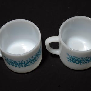 May include: Two white ceramic coffee cups with a teal floral design around the base. Each cup has a handle and is set against a black background. The cups are empty and appear to be vintage.