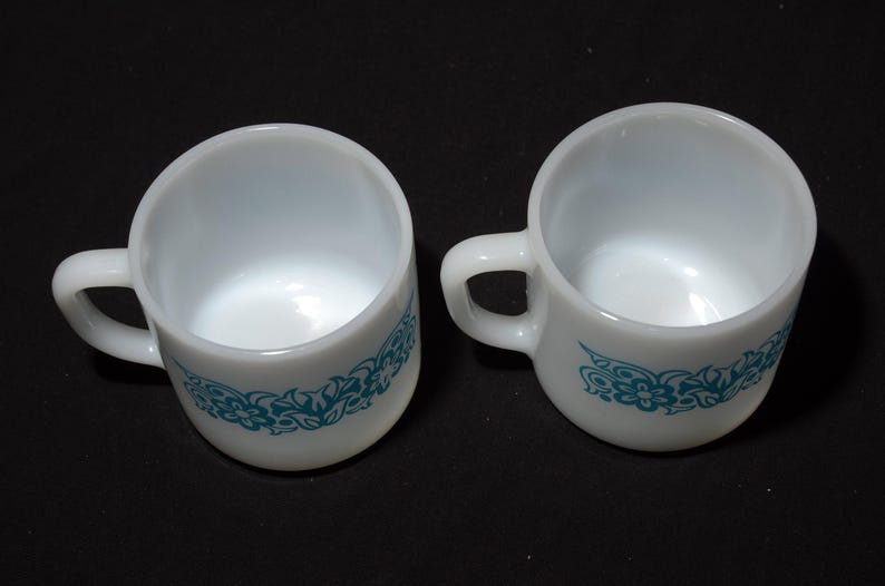 May include: Two white ceramic coffee cups with a teal floral design around the base. Each cup has a handle and is set against a black background. The cups are empty and appear to be vintage.
