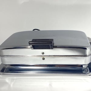 Puede incluir: Una gofrera rectangular de cromo con un acabado metálico pulido. El aparato cuenta con un asa de plástico negro en la tapa frontal y asas laterales de plástico negro. Dos pequeños tornillos son visibles en el borde frontal de la carcasa.