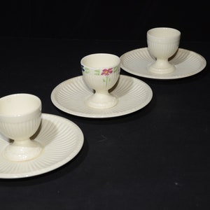 May include: Three white ceramic egg cups with floral designs on the side, each sitting on a white saucer with a fluted edge.