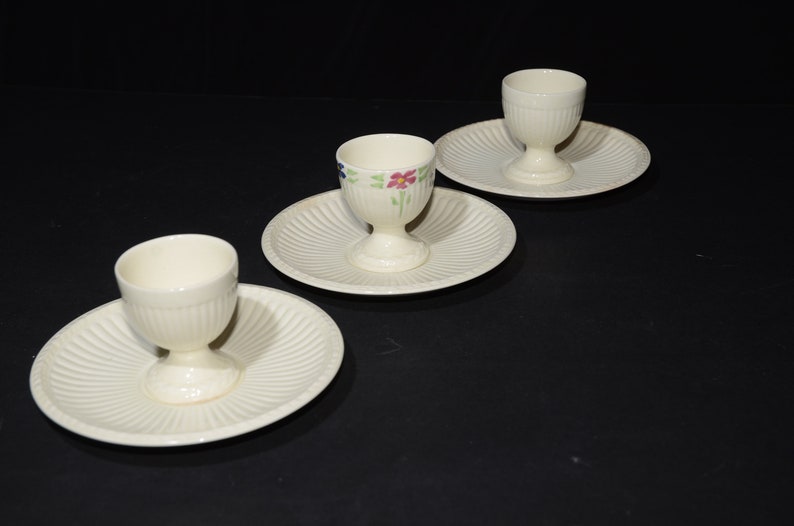 May include: Three white ceramic egg cups with floral designs on the side, each sitting on a white saucer with a fluted edge.