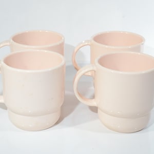 Può includere: Set di quattro tazze in ceramica rosa pallido con un design impilato. Le tazze hanno una forma arrotondata e un manico sul lato.