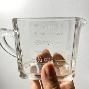 May include: A clear glass measuring cup with a handle and a pouring spout. The cup has measurements for 1/3 cup, 2/3 cup, and 1 cup. The cup is held by a person.