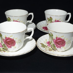 May include: A set of four white teacups and saucers, each decorated with a red rose and gold trim. The saucers have a scalloped edge. The teacups and saucers are a classic design for serving tea.
