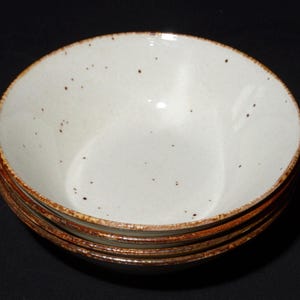 Puede incluir: Una pila de cuatro cuencos de cerámica blanquecina con superficies moteadas y un borde marrón rústico. Los cuencos están apilados uno encima del otro, mostrando su diseño simple pero elegante. Adecuado para servir sopas, ensaladas u otros platos.