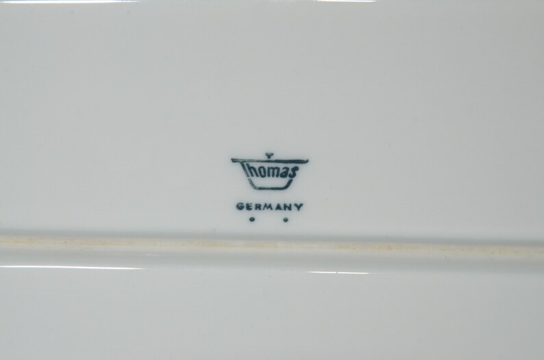 May include: A white rectangular ceramic serving platter with the Thomas Germany logo in dark blue. The logo includes the word "Thomas" above "GERMANY". The platter has a simple, elegant design.