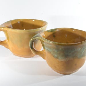 May include: Two ceramic mugs with a yellow glaze and green accents. The mugs have a unique, asymmetrical shape and a wide, rounded rim.