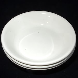 Op de afbeelding: Een stapel van drie witte keramische kommen. De kommen hebben een eenvoudig, klassiek ontwerp met een glad, wit interieur. De buitenkant heeft een subtiel randdetail. De kommen staan tegen een zwarte achtergrond.