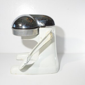 May include: A white and chrome manual citrus juicer with a lever on the side. The juicer is made of metal and has a chrome top.
