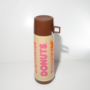 May include: A vintage Dunkin' Donuts thermos. The thermos has a brown lid and handle, with a tan body. The words "DONUTS" and "DUNKIN'" are printed in pink and orange. The thermos is designed to keep beverages hot or cold.