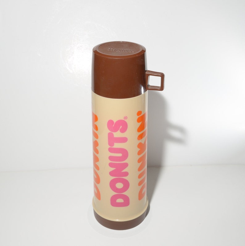 May include: A vintage Dunkin' Donuts thermos. The thermos has a brown lid and handle, with a tan body. The words "DONUTS" and "DUNKIN'" are printed in pink and orange. The thermos is designed to keep beverages hot or cold.