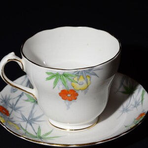 May include: White teacup and saucer set with a floral design. The cup and saucer feature a gold trim and a pattern of orange, yellow, and green flowers with blue and green leaves. The background is black.