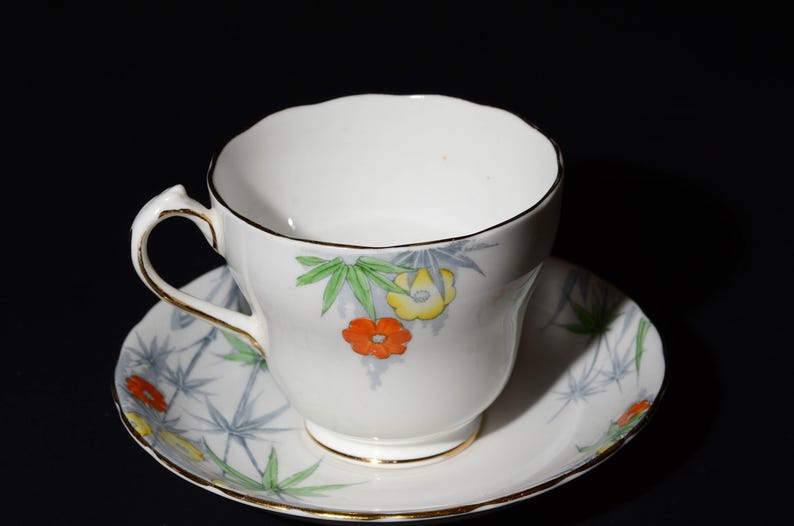 May include: White teacup and saucer set with a floral design. The cup and saucer feature a gold trim and a pattern of orange, yellow, and green flowers with blue and green leaves. The background is black.