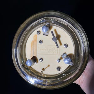 May include: A vintage, round clock with a cream-colored face and a gold-toned metal frame. The clock features silver-colored knobs and a small label with text. The clock's hands are visible, and the overall design suggests a classic aesthetic.