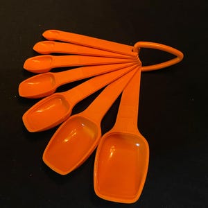 May include: A set of seven orange measuring spoons with a handle that connects them together. The spoons are made of plastic and are nested together.