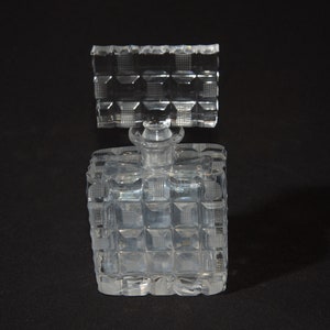 May include: A clear glass perfume bottle with a square body and a textured design. The bottle has a square stopper with a similar textured design.