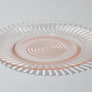 Puede incluir: Un plato de cristal rosa claro con un borde acanalado y un dise&ntilde;o de c&iacute;rculos conc&eacute;ntricos en el centro. El plato es redondo y proyecta una sombra sobre la superficie blanca.