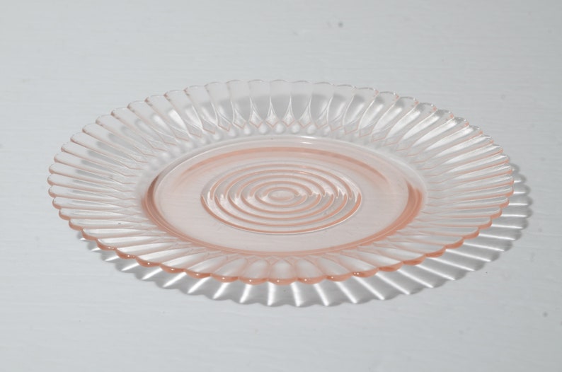 Puede incluir: Un plato de cristal rosa claro con un borde acanalado y un dise&ntilde;o de c&iacute;rculos conc&eacute;ntricos en el centro. El plato es redondo y proyecta una sombra sobre la superficie blanca.