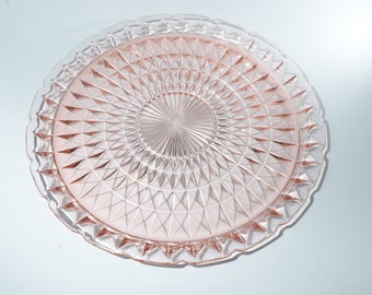 May include: A round, pink glass cake plate with a diamond pattern in the center and a scalloped edge. The plate is approximately 12 inches in diameter, perfect for serving desserts.