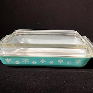 Puede incluir: Una fuente de horno rectangular Pyrex turquesa con tapa de cristal transparente. La fuente presenta un patrón de copos de nieve blancos alrededor de los lados. La tapa se asienta al ras en la parte superior de la fuente.