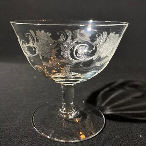 May include: A clear glass dessert dish with a delicate etched floral design. The dish has a wide, shallow bowl and a tall, slender stem.