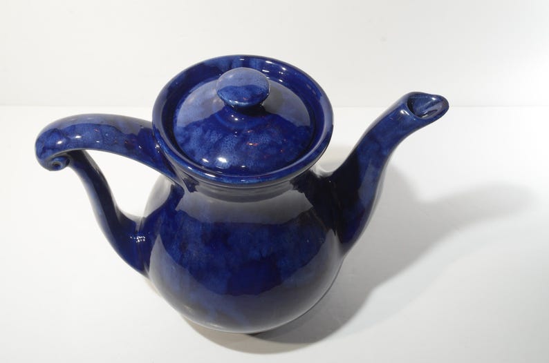 May include: A cobalt blue ceramic teapot with a rounded body and a curved spout. The teapot has a lid with a knob on top.