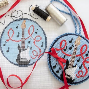 May include: A blue and white needlepoint ornament featuring a black electric guitar with red ribbon and musical notes. The ornament is unfinished and includes a blue cord for hanging.