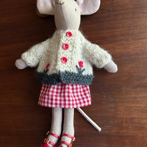 May include: A handmade mouse doll wearing a cream-colored knitted sweater with red flower accents and buttons, a red and white checkered skirt, and red leather shoes. The doll has large ears and a long tail.