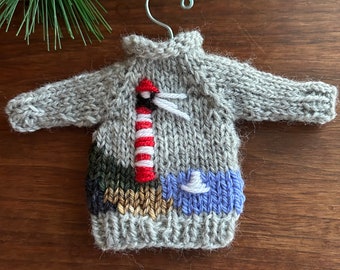 May include: A gray knitted sweater ornament with a lighthouse design. The lighthouse is red and white with a white hat on top. The sweater has a blue and white stripe on the bottom.