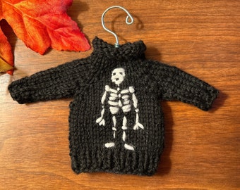 May include: A black knitted sweater with a white skeleton design. The sweater is hanging on a hanger.