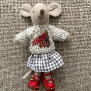 May include: A plush mouse toy with a beige fabric body, wearing a chunky knit sweater featuring a brown horse motif, a brown and white gingham skirt, and bright red leather-style Mary Jane shoes with gold-tone buckles and visible stitching.