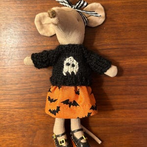 May include: A plush mouse doll wearing a black knit sweater with a white ghost graphic, an orange skirt featuring a black bat pattern, and black buckled shoes. The mouse has a black and white striped ribbon tied around its ears.