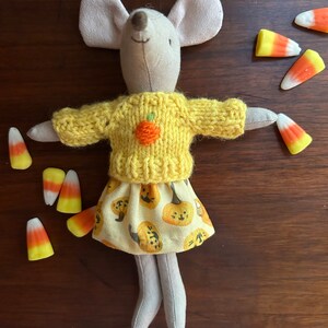 May include: A stuffed mouse doll wearing a yellow sweater with an orange pumpkin and a skirt with pumpkin designs. Candy corn is scattered around the doll on a dark brown wooden surface. The doll has gray ears and legs.