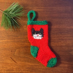 May include: A small, hand-knitted Christmas stocking in red with green accents. The stocking features a black and white cat face design. A green loop is at the top for hanging. A sprig of pine needles is in the background.
