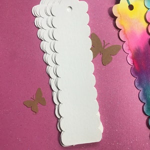 May include: A stack of white scalloped edge paper tags with a hole punched at the top. The tags are on a pink sparkly background.