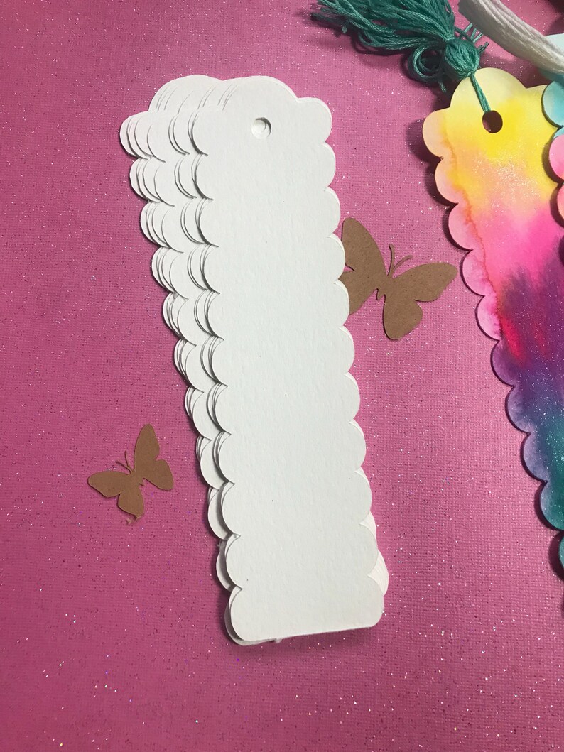 May include: A stack of white scalloped edge paper tags with a hole punched at the top. The tags are on a pink sparkly background.