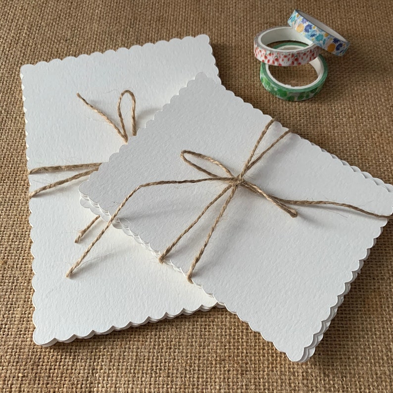 May include: Two white cards with scalloped edges are tied together with twine. The cards are stacked on top of each other and the twine is tied in a bow.