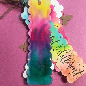 May include: A set of colorful watercolor bookmarks with scalloped edges. The bookmarks feature a variety of colors, including pink, blue, green, and yellow. The text on one bookmark reads "To be different".