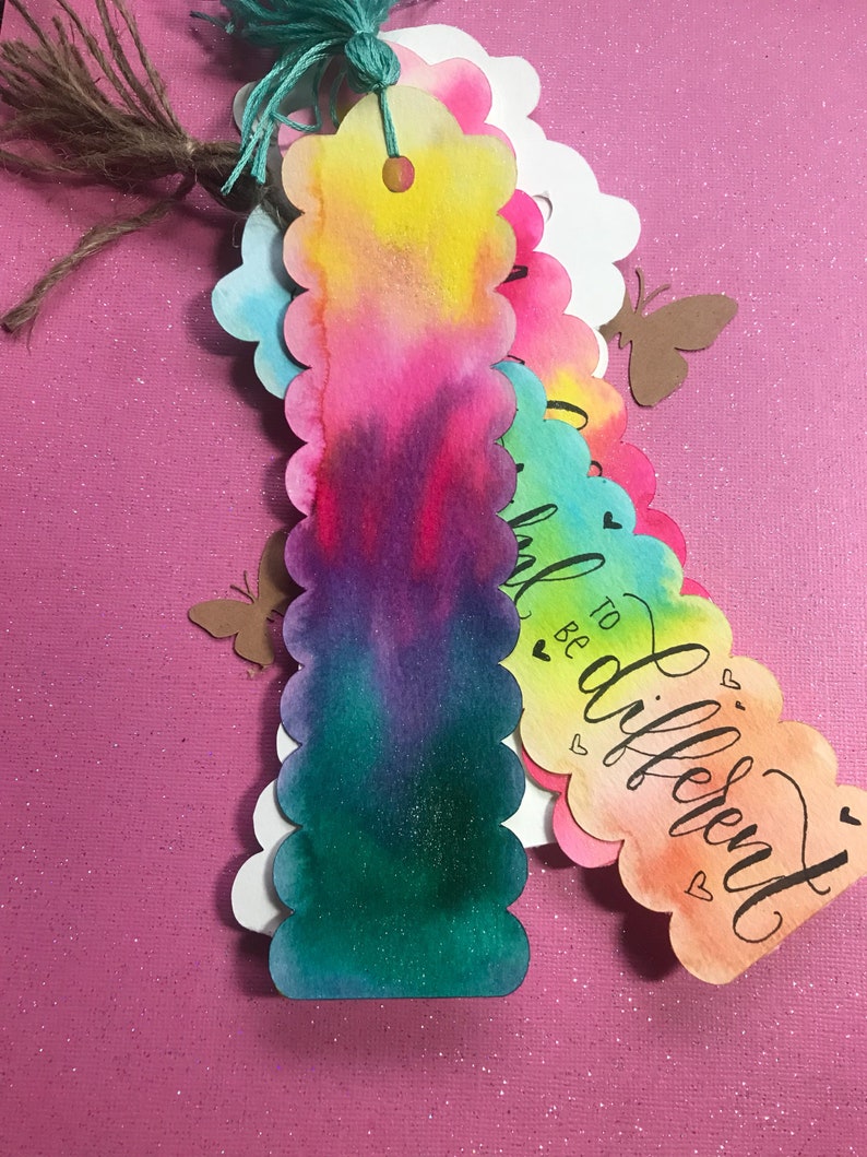 May include: A set of colorful watercolor bookmarks with scalloped edges. The bookmarks feature a variety of colors, including pink, blue, green, and yellow. The text on one bookmark reads "To be different".