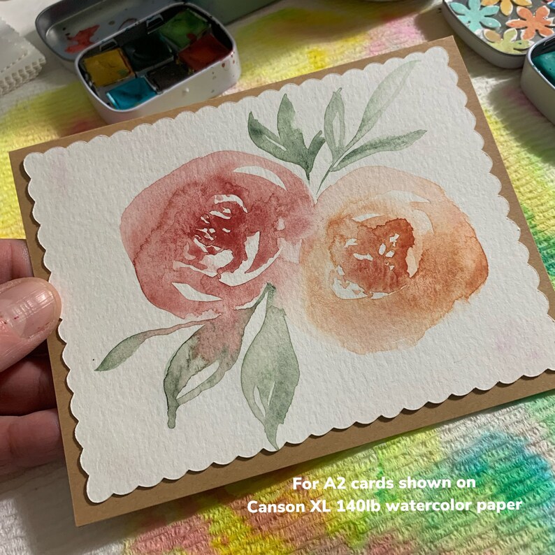 May include: A watercolor painting of two pink and orange flowers with green leaves on a white card. The card is on a brown background with the text "For A2 cards shown on Canson XL 140lb watercolor paper".