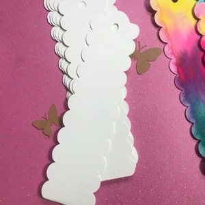 May include: A stack of white scalloped edge paper tags with a hole punched at the top. The tags are on a pink sparkly background.