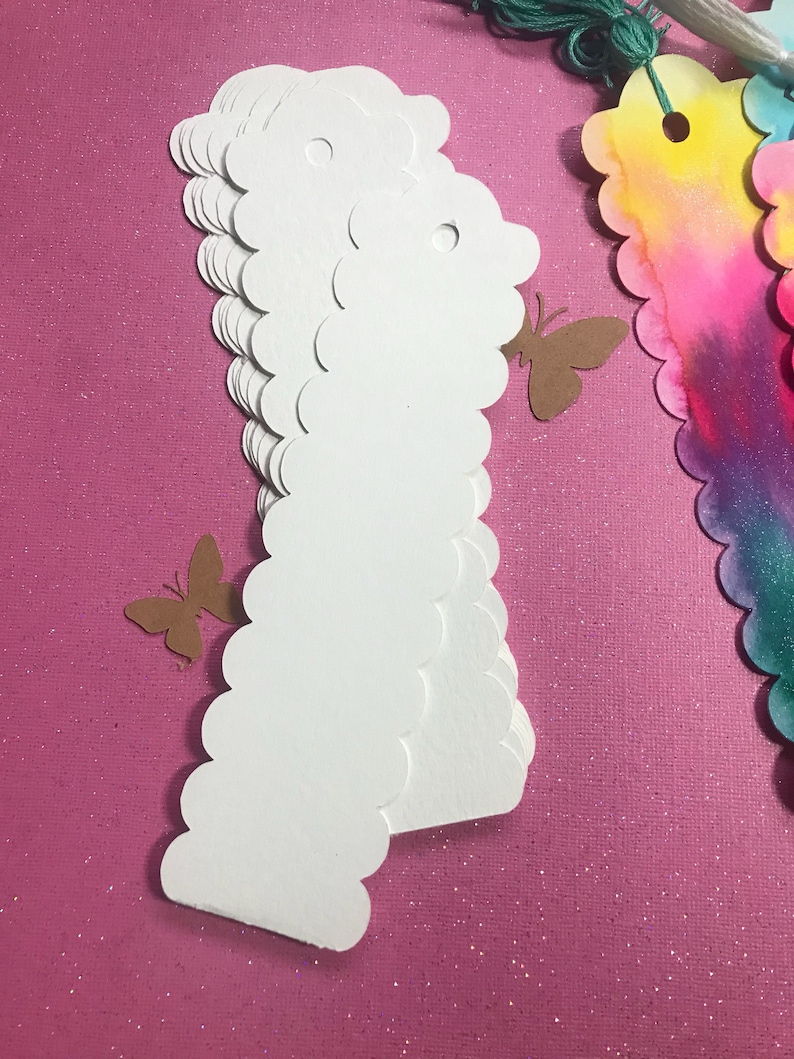 May include: A stack of white scalloped edge paper tags with a hole punched at the top. The tags are on a pink sparkly background.