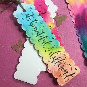 May include: A colorful watercolor bookmark with scalloped edges and the text "It's beautiful to be different". The bookmark has a rainbow gradient of colors, including pink, orange, yellow, green, and blue. There are small hearts and butterflies on the bookmark.